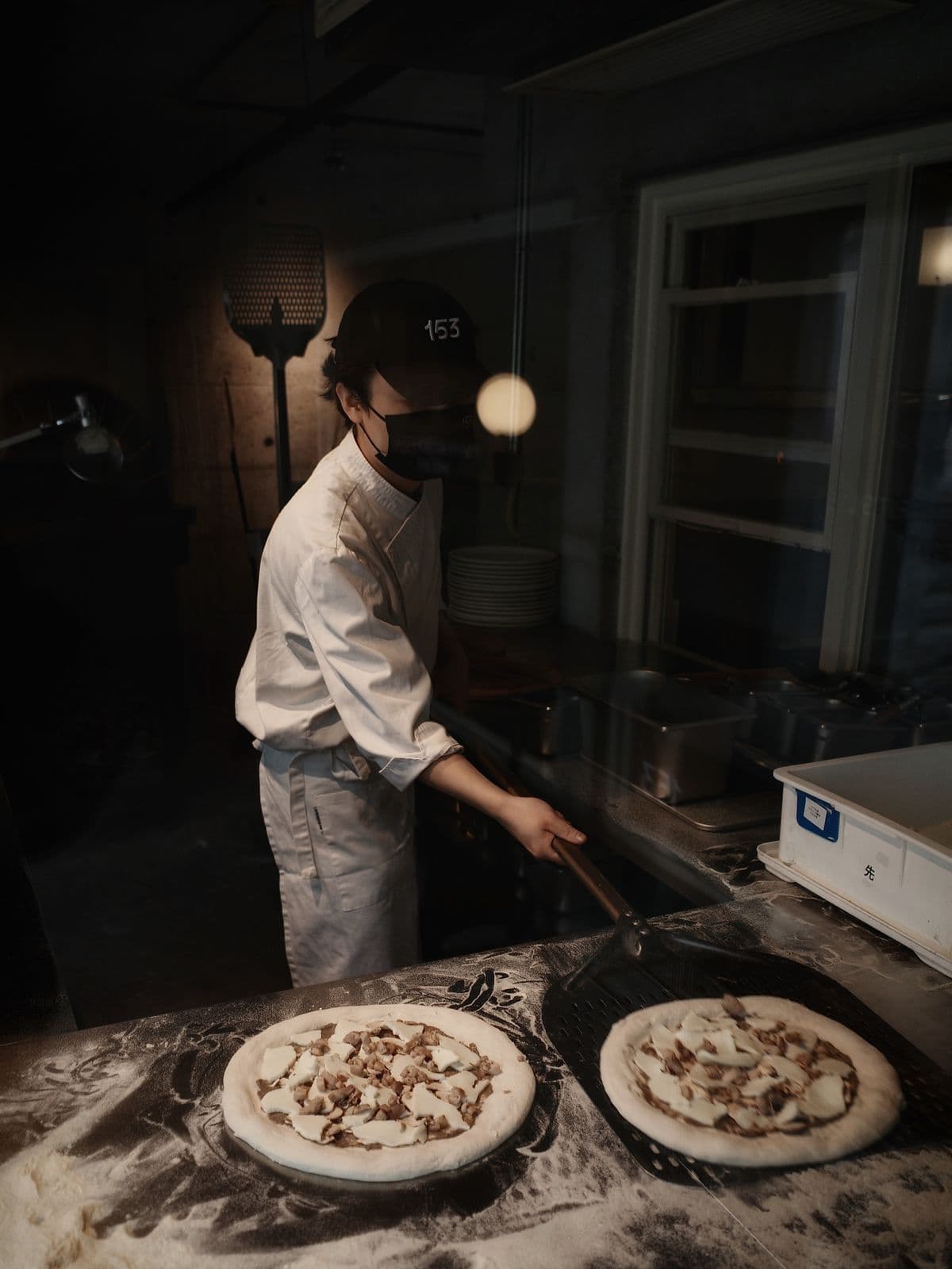 A pizzaiolo sliding two pizzas into a wood-fired oven in a dark kitchen