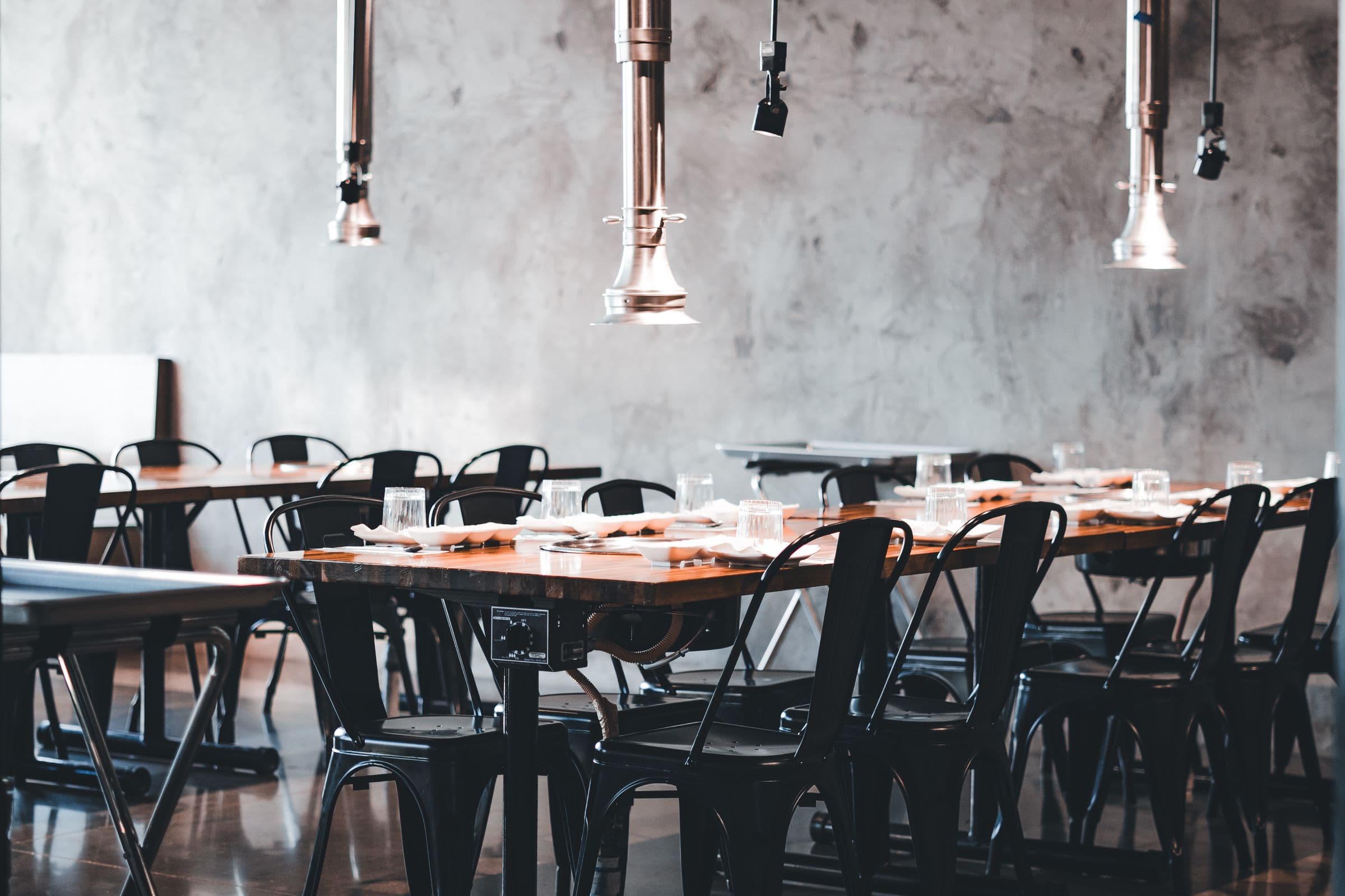 The dining room: raw concrete wall, steel pendants, laid tables in black bentwood chairs